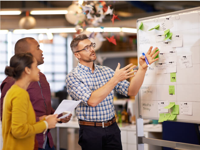 Un groupe de personnes debout autour d'un tableau blanc écoutant un homme expliquer ce qui est sur le tableau